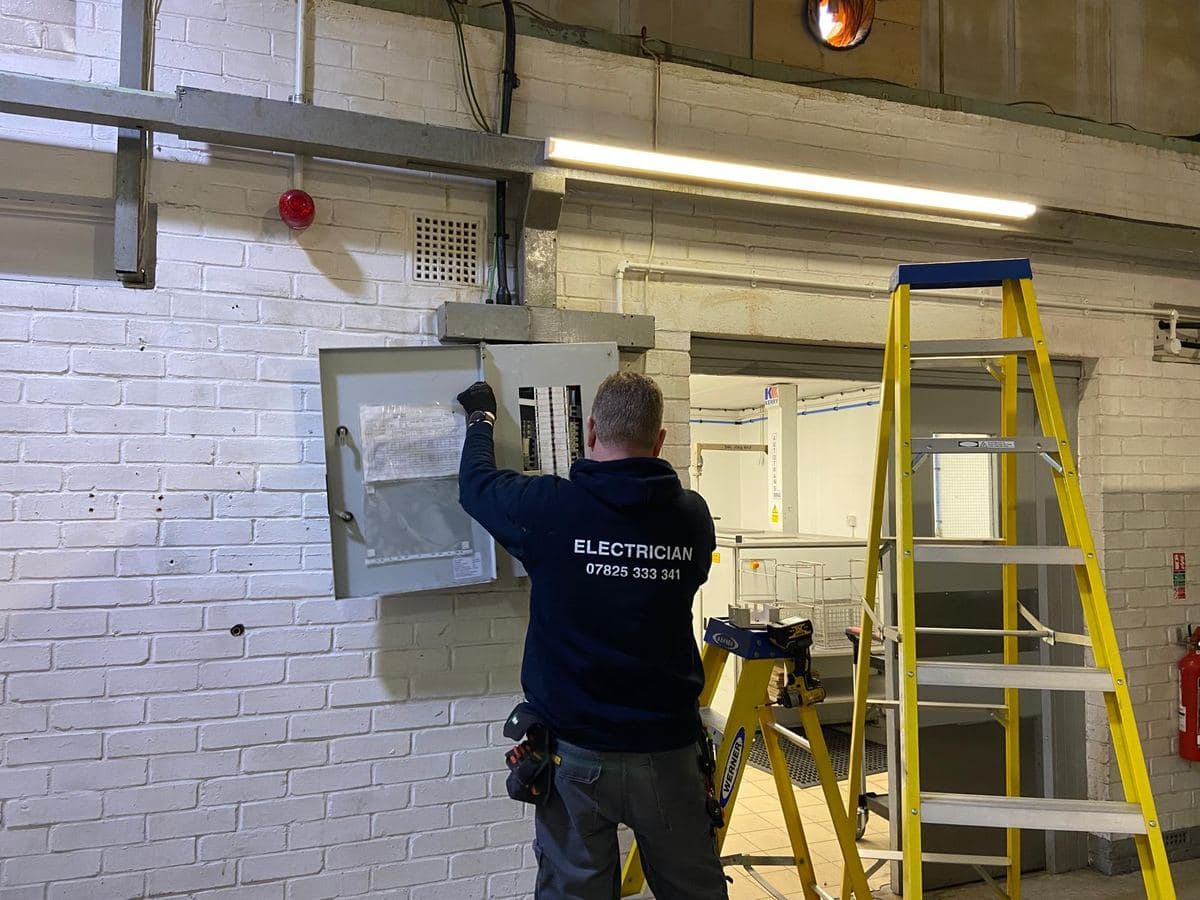 Electrician working at a distribution board on a factory wall, step ladders beside